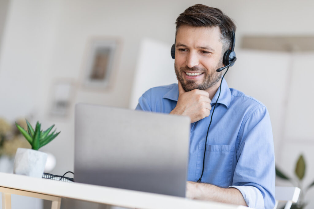 Hotline Support Service. Handsome male call center operator in headset working with laptop in modern office, looking at computer screen, free space